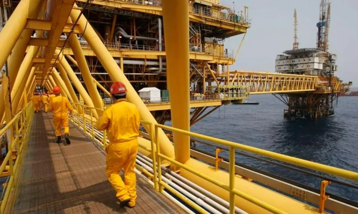 Workers in yellow coveralls and red helmets walk along a catwalk on an offshore oil platform overlooking the sea.