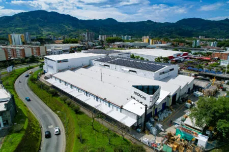 Aerial view of a manufacturing campus with white buildings, Hitachi signage, nearby roads, and green hills in the distance.