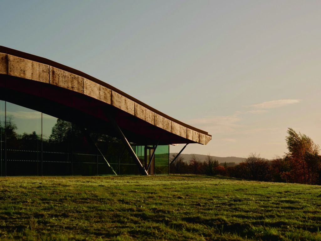 - Petróleo y Energía Curved, stone-clad overhang of a modern building with glass walls, set on a grassy landscape at dusk.