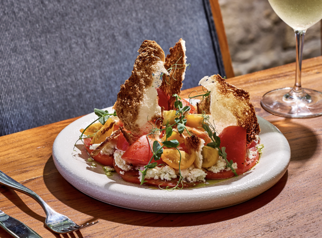 QUINTO RESTAURANTE MIAMI 1 - Petróleo y Energía Plate of tomato, cheese and herb salad with crispy bread shards on a white plate; wine glass on wooden table in the background.