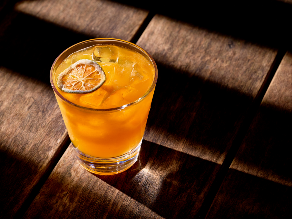 Quinto -Miami Spice 2026 - Caipi Maracuyá - Petróleo y Energía Orange juice with ice and a lemon slice garnish in a short glass on a sunlit wooden table.