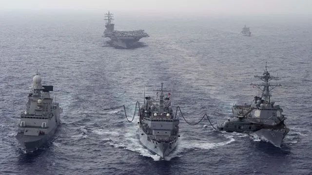 Four gray naval ships sail in formation on a calm sea; three foreground ships are connected by cables, with a large aircraft carrier visible in the hazy distance.