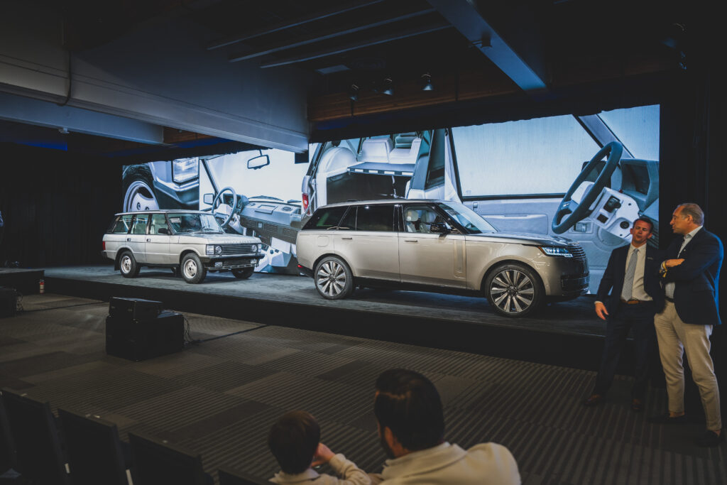 RANGE ROVER MATCHED PAIR ACHIEVES RECORD_RM SOTHEBYS MONTEREY AUCTION_210826_02 - Petróleo y Energía Display stage with multiple cars showcased indoors, large screen backdrop showing car interiors, and two men in suits conversing on the right.
