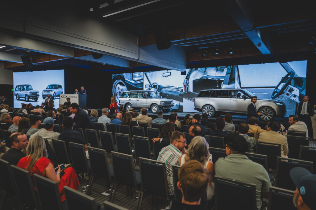 RANGE ROVER MATCHED PAIR ACHIEVES RECORD_RM SOTHEBYS MONTEREY AUCTION_210826_03 - Petróleo y Energía Audience seated in a conference hall watching a car presentation with large screens showing vehicles on stage behind the speakers.