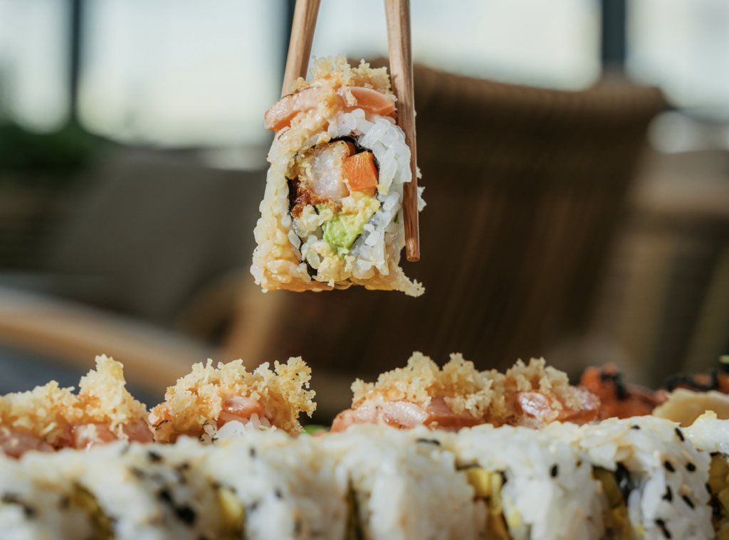 SUGAR RESTAURANTE MIAMI 1 - Petróleo y Energía Close-up of a sushi roll held by chopsticks, with tempura crunch, avocado, and fish inside.