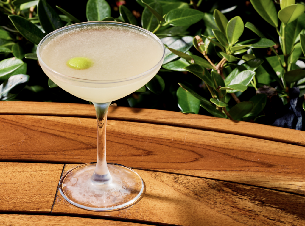 SUGAR RESTAURANTE MIAMI 2 - Petróleo y Energía Cocktail glass with pale yellow drink and a green olive on a wooden table, outdoor greenery in the background