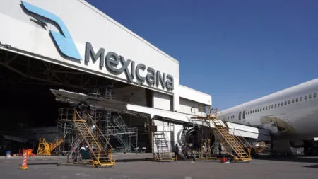 Ground crew work on a large Mexicana airliner parked at a maintenance hangar with scaffolding and mobile stairs nearby.
