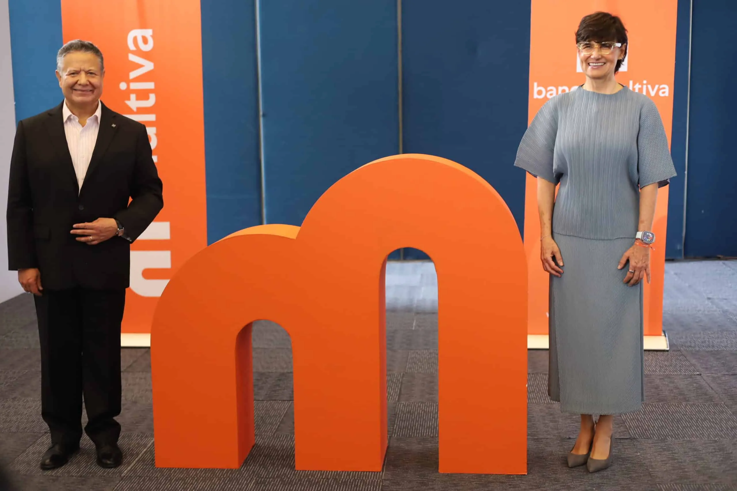 Man in a black suit and a woman in a gray dress stand beside large orange abstract shapes in a blue exhibition backdrop.