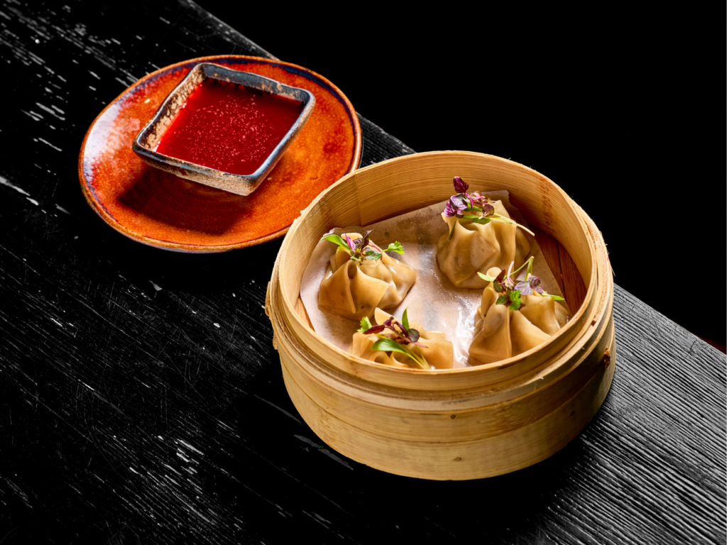 Tea Room -Miami Spice 2026 - Golden Siu Mai - Petróleo y Energía Dim sum dumplings in a bamboo steamer with microgreens, beside a square red sauce dish on a dark wooden table