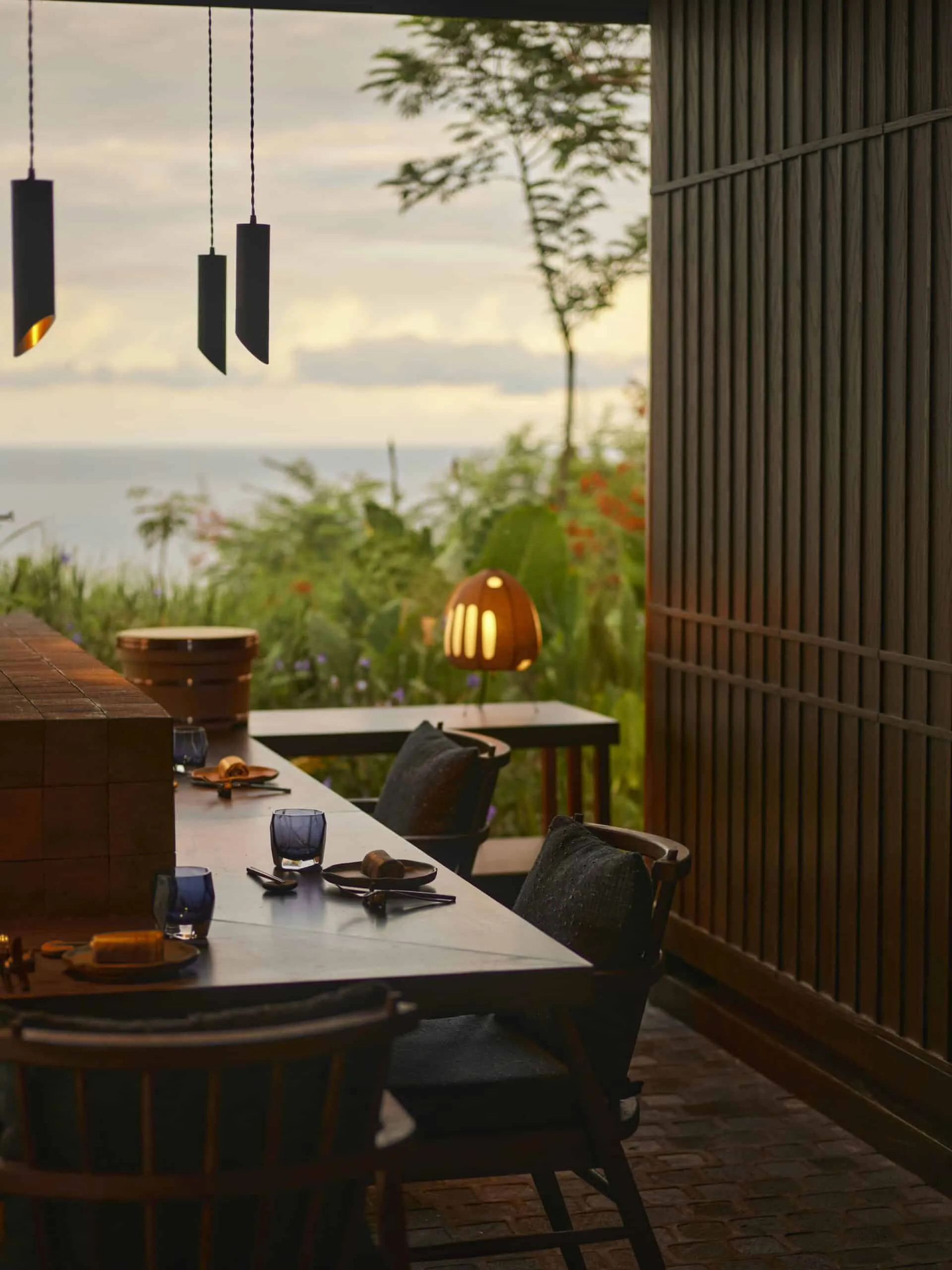 Outdoor dining table set for an evening meal with blue glasses, in a warm-lit patio overlooking a garden and ocean horizon at sunset.