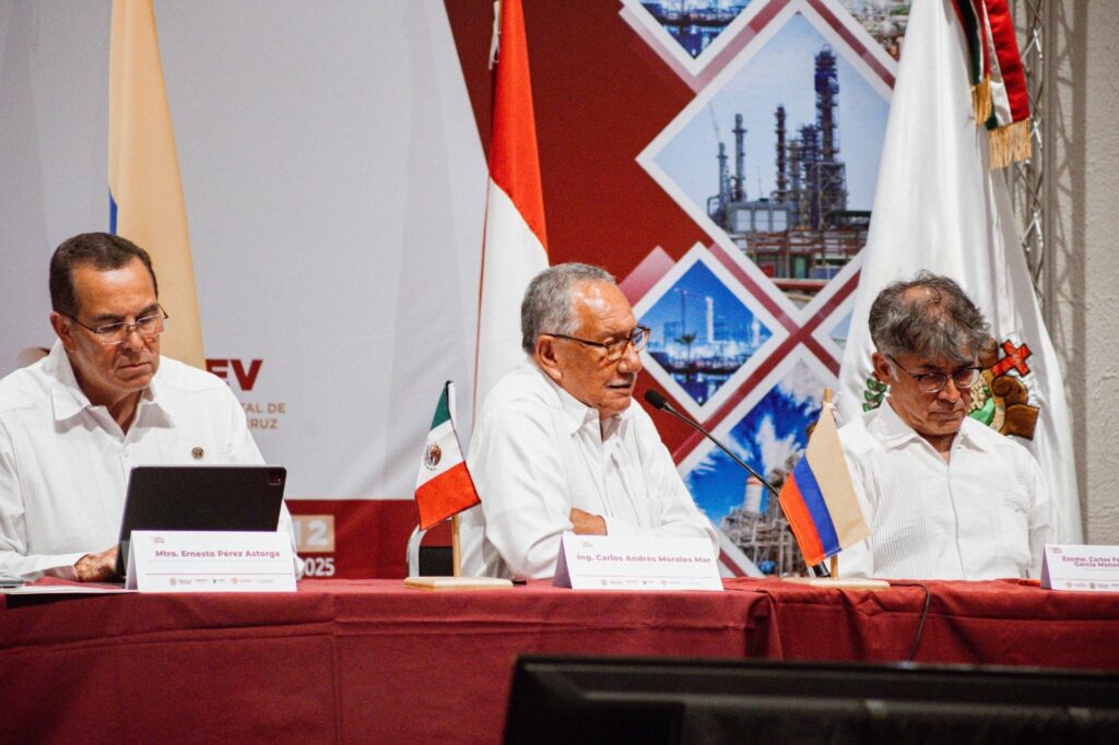 WhatsApp Image 2026-08-12 at 44034 PM - Petróleo y Energía Panel of three men in white shirts at a conference, with flags (Mexican and another) on the table and a red backdrop displaying industrial imagery behind them.
