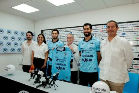 Group of six people at a press conference, two players in blue jerseys holding a matching kit in front of sponsor backdrop with logos, microphones on the table in front of them.