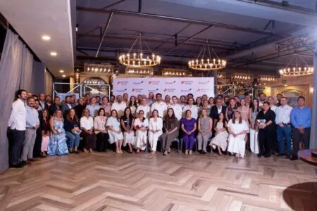 A large group of professionals posing for a formal group photo indoors, with a banner backdrop and chandeliers above.
