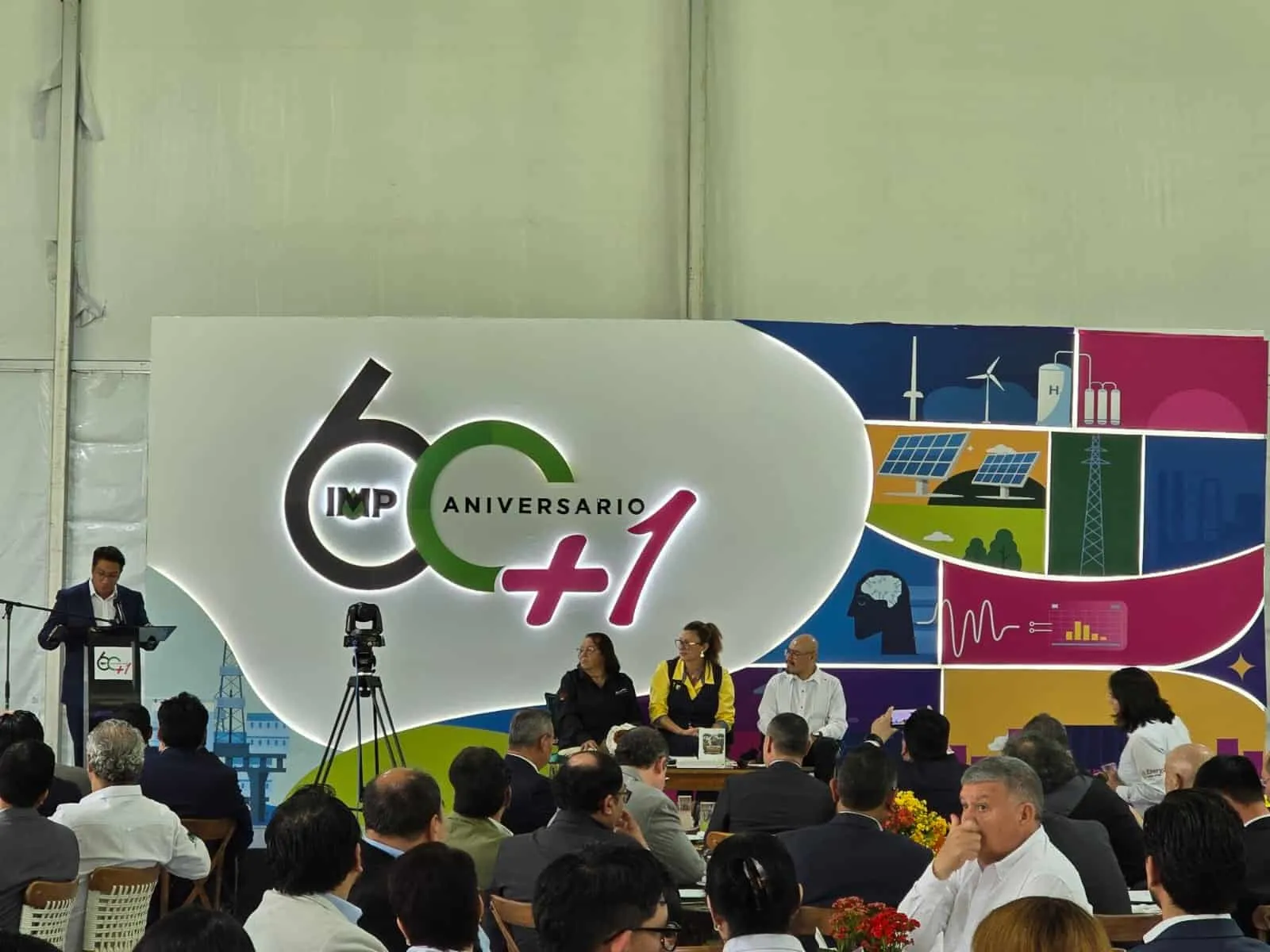 Man at podium speaking to an audience at a 60th anniversary event with a large colorful stage banner behind him and seated panelists nearby.