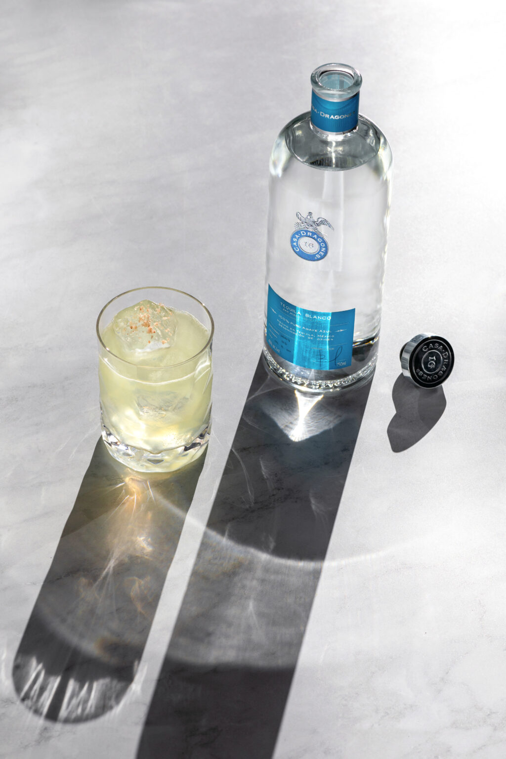 a-more-mexican-margarita - Petróleo y Energía Casa Dragones tequila bottle with blue label beside a glass of pale drink on a gray surface with long diagonal shadows.