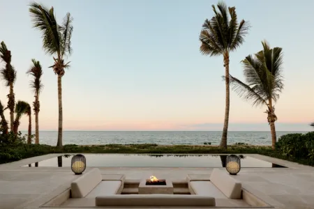 Ocean view at sunset with tall palm trees, calm sea, and a modern poolside lounge with a fire pit in the foreground.