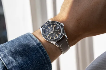 Wristwatch with a dark dial and beige markers worn on a forearm, denim sleeve visible nearby.