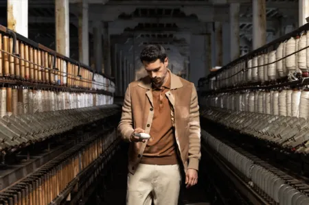 Man in a tan coat walks between rows of yarn-spinning machines in a dim textile mill, inspecting a small object in his hand.
