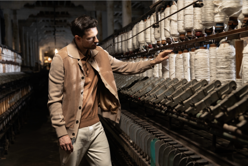 calderoni 2 - Petróleo y Energía Man in a beige coat touches a row of white thread spools in a dim textile factory aisle.