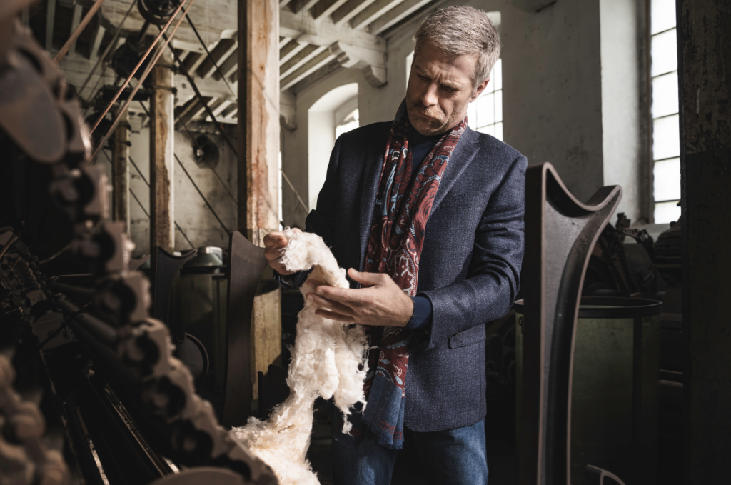 calderoni 5 - Petróleo y Energía Man in a blue blazer and scarf examines a fluffy bundle of wool in a dusty, industrial workshop filled with machines and wooden beams