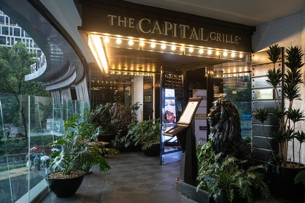 capitalre - Petróleo y Energía Entrance to The Capital Grille with two lion statues and lush plants under warm marquee lights behind a glass facade.
