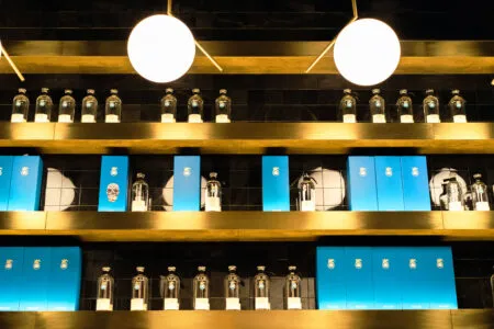 Row of clear glass bottles on a shelf with blue boxes beneath, against a dark tiled wall and warm lighting.