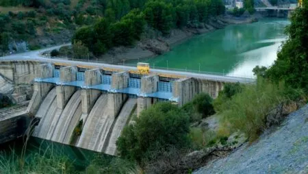 Dam with blue spillways across a green river; a yellow maintenance vehicle sits on the dam crest along a road.