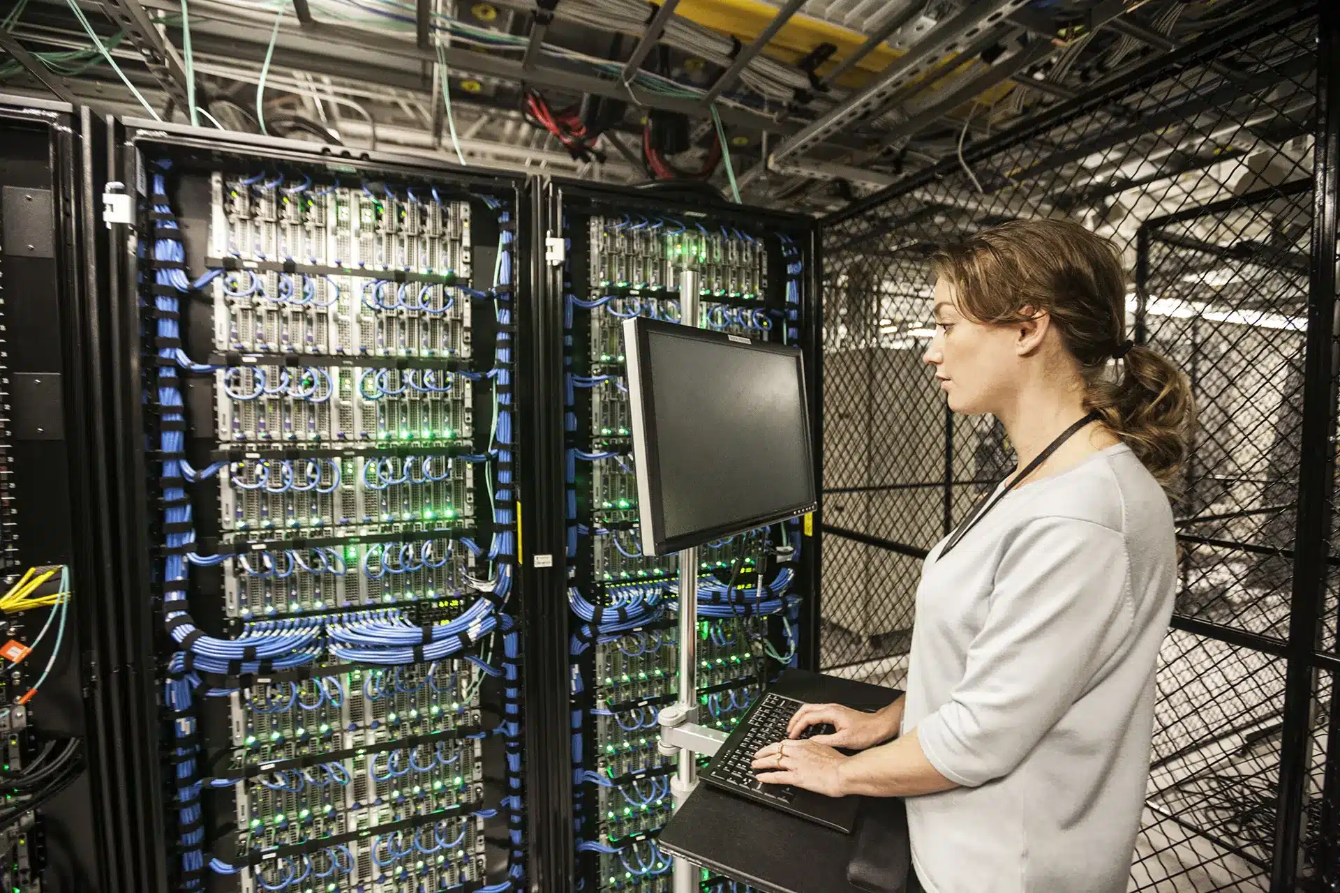 Female technician working at a server rack, inspecting and typing on a keyboard mounted on a sliding tray amid blue network cables and illuminated green status LEDs.