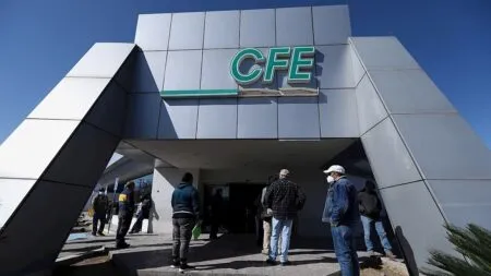 People wearing masks stand in a queue outside a modern CFE building with a green sign under a clear blue sky.