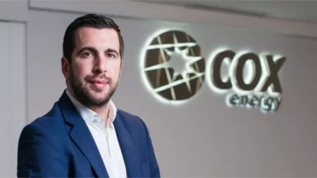 Man in a blue suit standing in front of a illuminated COX energy sign in a corporate lobby.