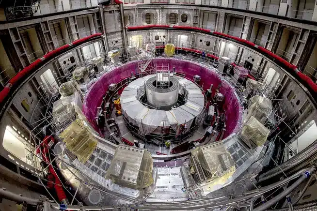 Interior of a large scientific reactor/particle accelerator chamber, with a central cylindrical core surrounded by a purple circular ring and metal scaffolding.