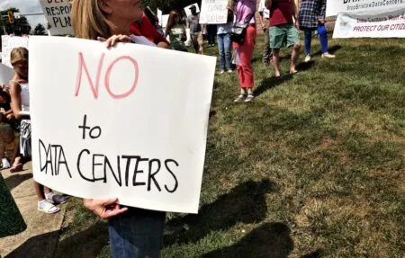 Person holding a large sign that says 'NO to DATA CENTERS' at an outdoor protest on a grassy area