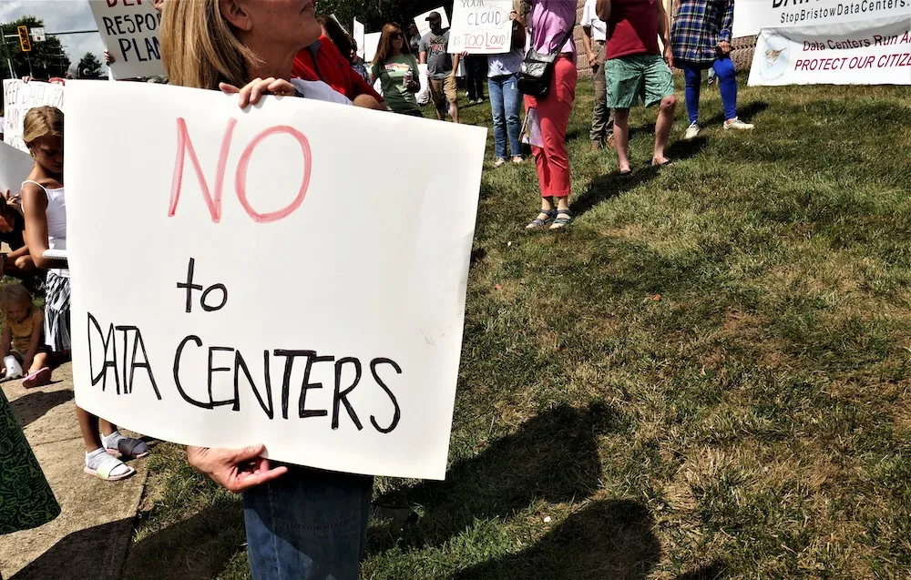 Person holding a large sign that says 'NO to DATA CENTERS' at an outdoor protest on a grassy area