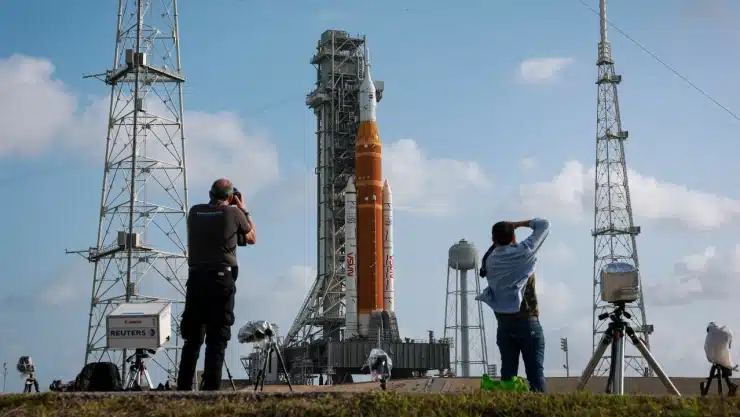 Rocket on a launch pad with photographers taking photos and tall support towers in the background.