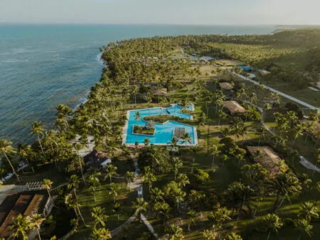Aerial view of a tropical resort with a turquoise pool, palm trees, and scattered bungalows along the coastline. The ocean is to the left.