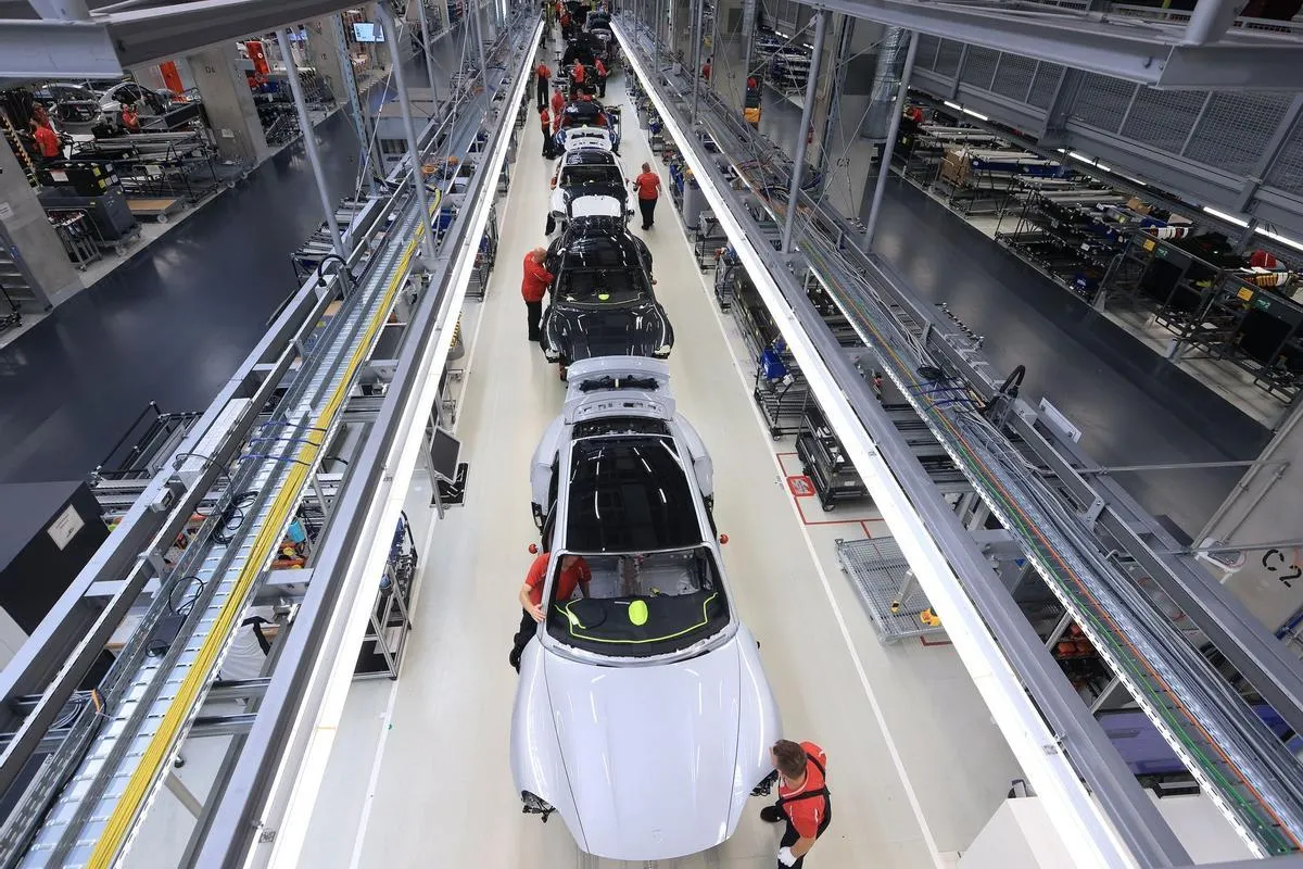 Automobile assembly line with white and black cars in a multi‑level production hall and workers in red shirts assembling vehicles along a conveyor track.