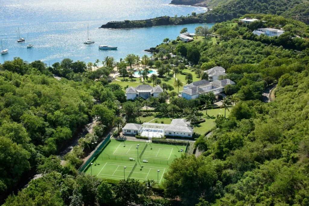 Aerial view of a tropical resort: tennis courts in the foreground, white villas, palm trees, and a turquoise bay with boats.