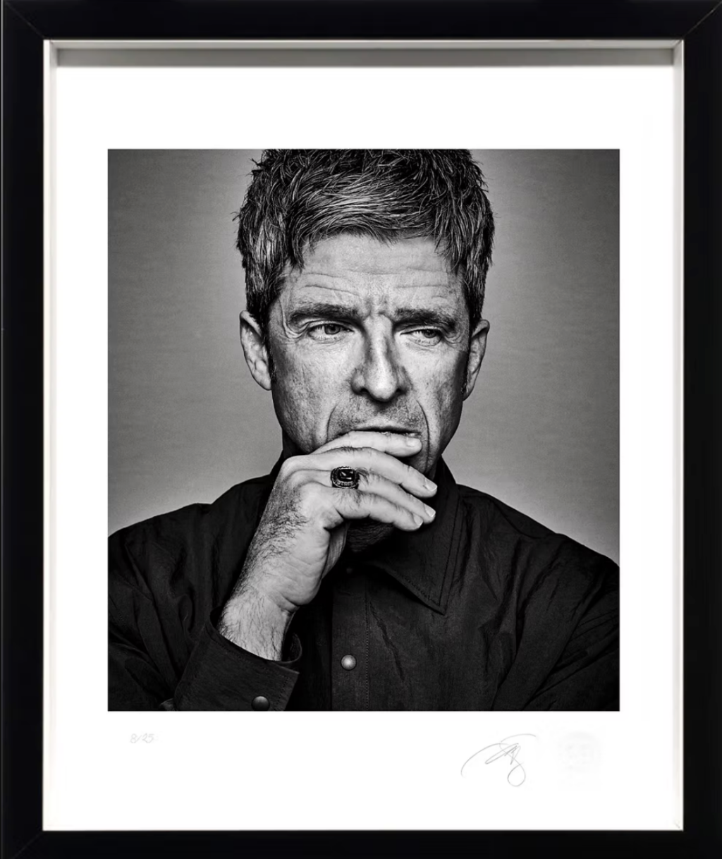 El cantante Noel Gallagher - Petróleo y Energía Black-and-white framed portrait of a thoughtful man with short hair, resting his chin on his hand and wearing a dark button-up shirt and ring on his finger.