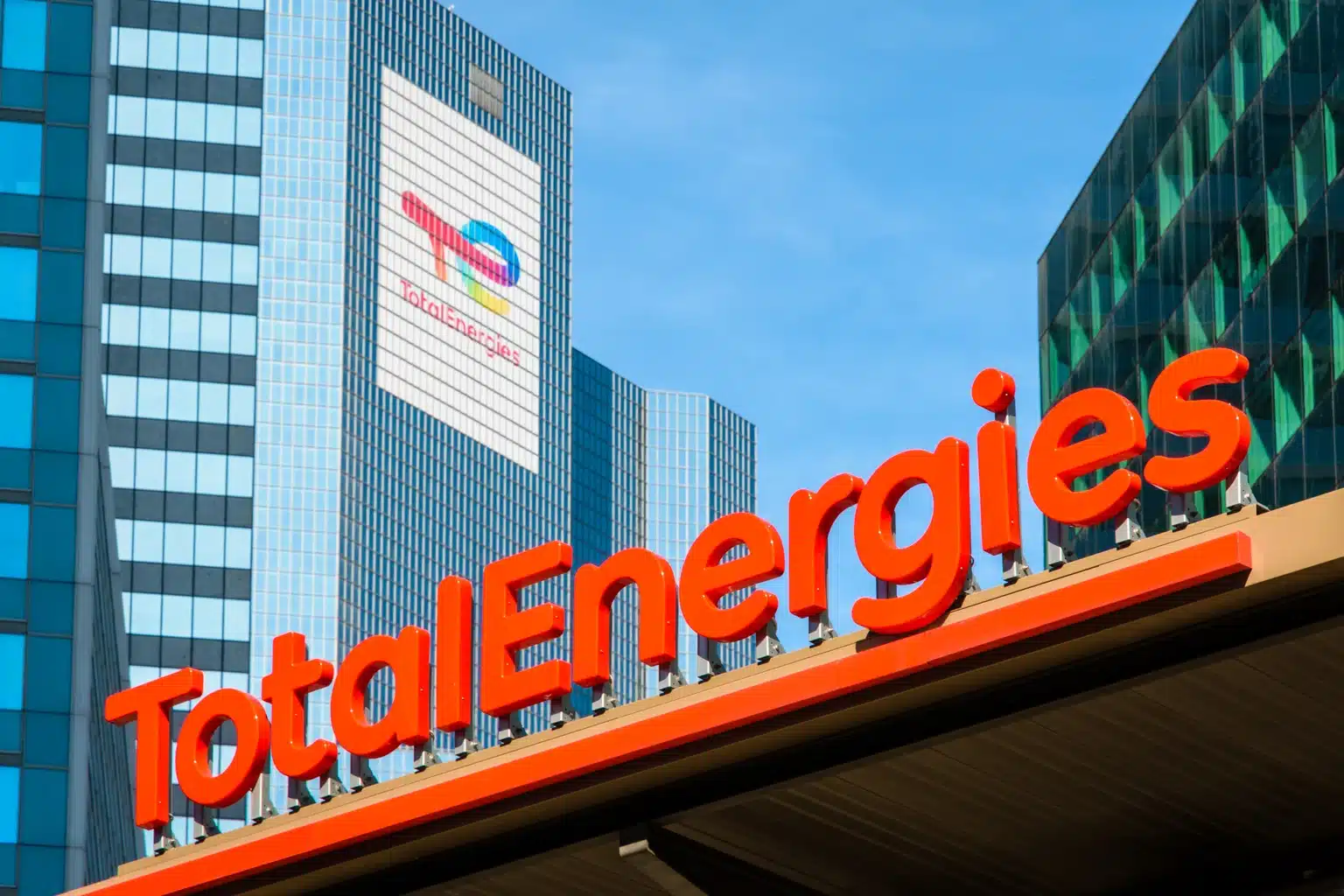 Red TotalEnergies sign on a modern building facade against a blue sky.