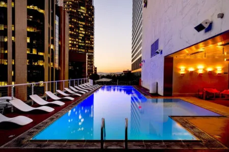 Rooftop hotel pool at sunset with city skyscrapers, white lounge chairs along the poolside.