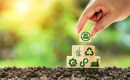 A hand places a 'Net Zero' block atop a stack of wooden blocks with sustainability icons, on soil with a green blurred background.
