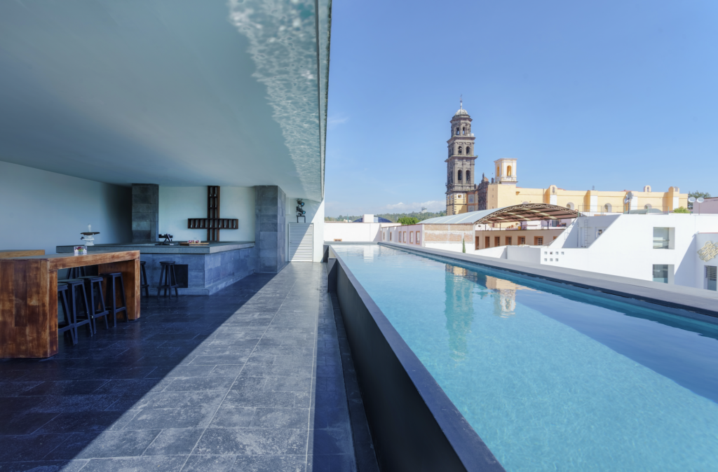 la purificadora 1 - Petróleo y Energía Rooftop pool terrace with a long blue pool on the right, a wooden bar on the left, and a historic bell tower visible in the distance under a clear sky.