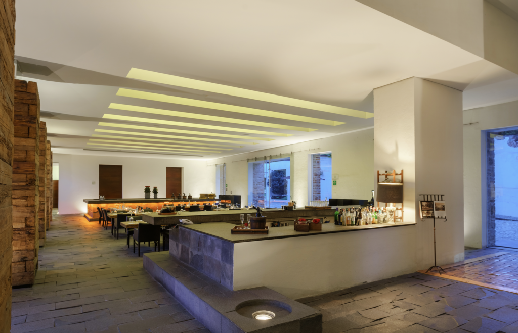 la purificadora 10 - Petróleo y Energía Spacious bar area with a long central counter, bottles and glasses, and yellow lighted ceiling slats; stone floor and brick wall on the left.