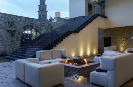 Outdoor lounge area with white sectional couches around a rectangular fire pit, stone walls, and a modern staircase-lit ambiance at dusk/evening.