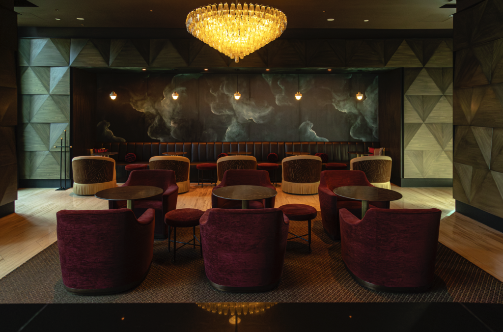 llobsiadian_sofitel 3 - Petróleo y Energía Luxurious lounge with red velvet chairs, round tables, and a large crystal chandelier overhead.