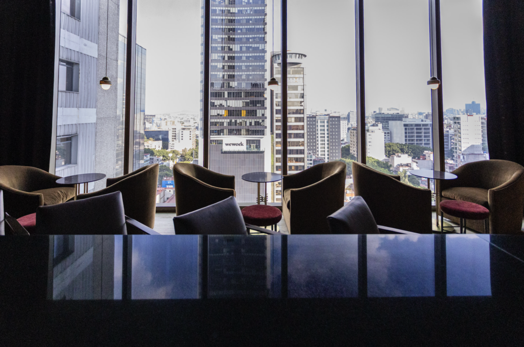 - Petróleo y Energía Cozy modern lounge with dark chairs and small round tables facing a city skyline through tall windows.