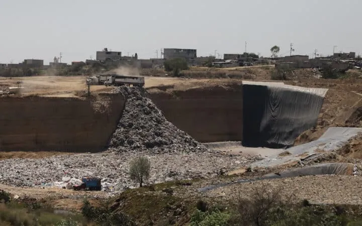 Mining site with a large rock pile beside a black-lined retaining wall; dusty terrain and distant buildings on the horizon.