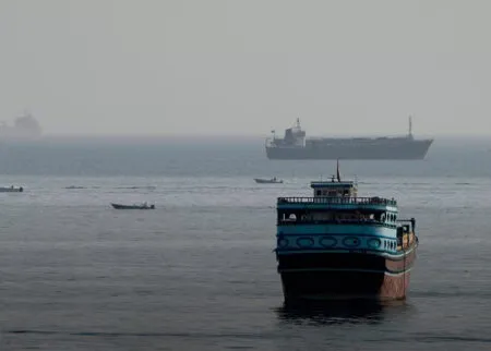 Blue and black passenger ferry in the foreground on calm water, with a large cargo ship in the hazy distance.