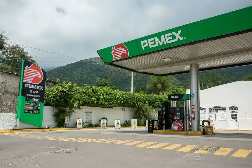 Gas station with a green PEMEX canopy and pumps, set against a hillside landscape and cloudy sky.