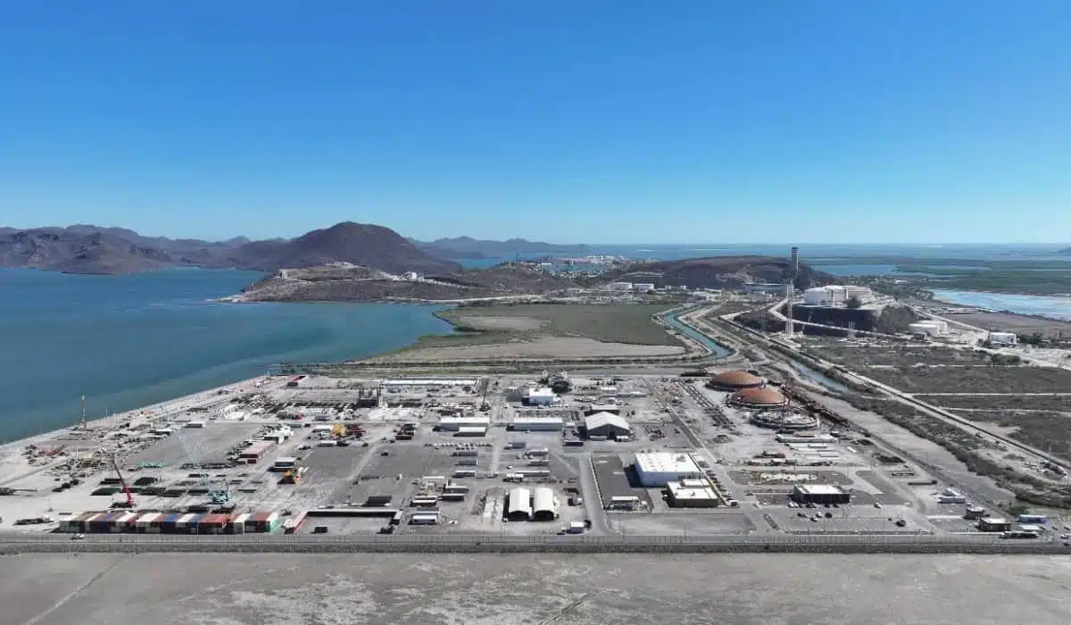 A coastal industrial port with container yards, warehouses, and paved lots viewed from above, with blue water and distant hills in the background.